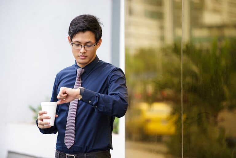 Un homme brun portant des lunettes; Il tient un café dans sa main et regarde sa montre de l'autre main